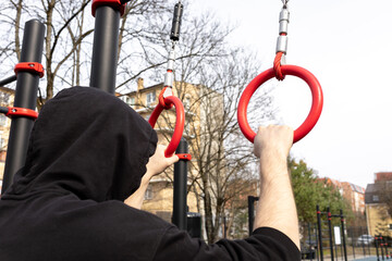 A man in a hoodie holds onto gymnastic rings located on an outdoor sports ground.