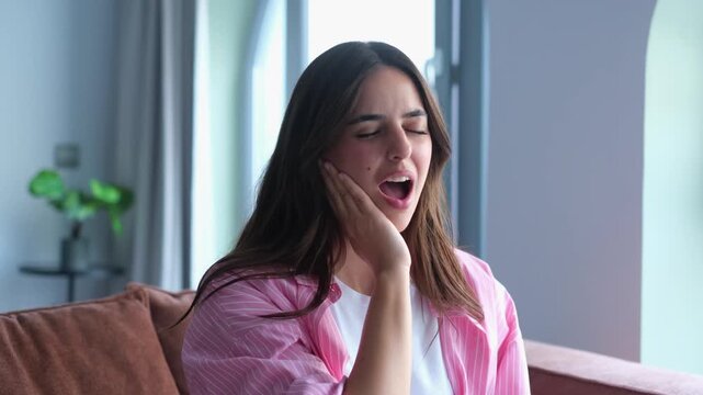 A young woman feels discomfort in her jaw while yawning. She rubs her face, indicating potential pain or an issue with her temporomandibular joint, possibly due to tiredness or dental problems.