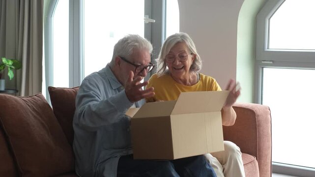 A loving senior couple sits on their living room couch, eagerly opening a brown cardboard box. Their faces light up with joy and surprise as they discover the unexpected contents