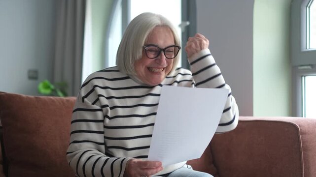 A joyful elderly woman on a cozy sofa happily reads an important document, reacting with surprise, a big smile, and excited laughter. She clenches her fist in triumph, celebrating wonderful good news 