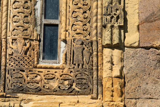 Gergeti trinity church's ancient stone facade features a carved window framed by detailed biblical scenes, warriors and traditional orthodox motifs, weathered but intricate