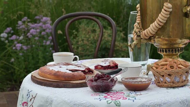 A rustic tea setting with golden samovar, fresh strawberries, jam, pie, and bagels arranged on an embroidered tablecloth outdoors.