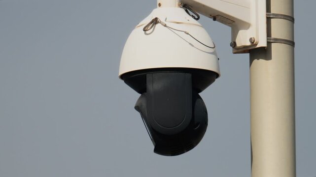 White outdoor surveillance dome camera rotates slowly monitoring public space against gray sky background.