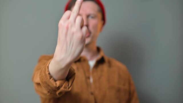 A man wearing a brown shirt and red beanie makes a hand gesture, displaying a bold expression.