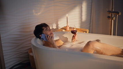 Bath girl calling mobile phone lying foam water with red wine glass evening spa
