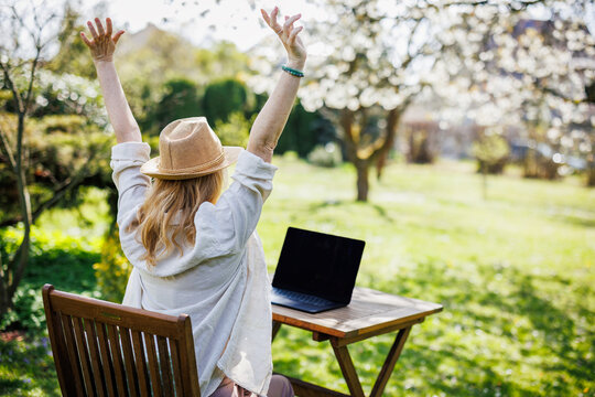 Woman stretching arms while working on laptop in spring garden. Stress relief. Freelancer taking break during administrative remote work