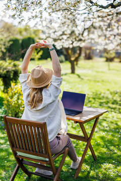 Woman stretching hands while working with laptop outdoors. Stress relief. Exercising body during remote work break in spring garden