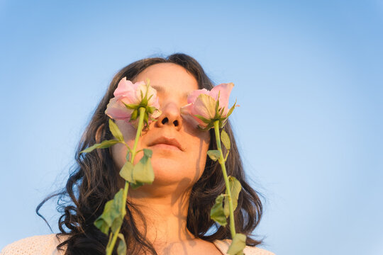 young millennial woman with flowers in front of eyes against blue sky, holding roses , has spring mood, enjoys pleasant smell in spring