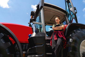 Naklejka premium Female farmer standing with crossed arms by tractor outdoors, low angle view.