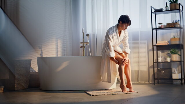 Girl applying legs cream sitting bathtub home. Resting woman rubbing moisturizer