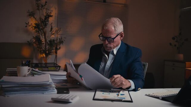 Mature overworked entrepreneur feeling stressed while analyzing business reports and paperwork, working late in a dark office