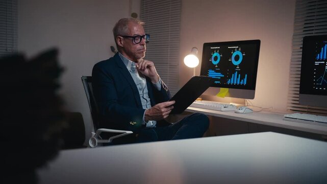 Concentrated senior businessman in a suit working overtime, reviewing company reports and comparing data with charts on his computer screen