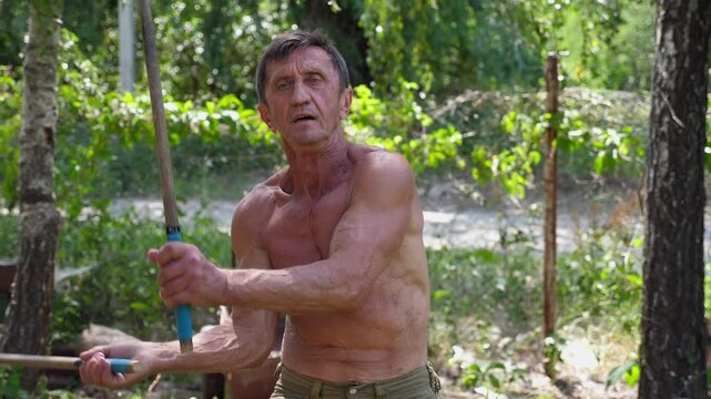 Adult male athlete showing fighting techniques with wooden sticks outdoor. Older sportsman demonstrating wing chun kung fu. Old man exercising martial arts at summer day. Concept of healthy lifestyle