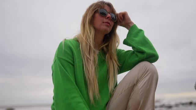 Stylish woman in green sweater poses outdoors on a cloudy day.