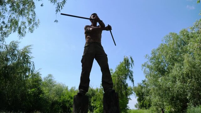 Old trainer standing on high stumps and demonstrating wing chun kung fu. Adult male athlete showing fighting techniques with wooden sticks outdoor. Older man exercising martial arts at summer day
