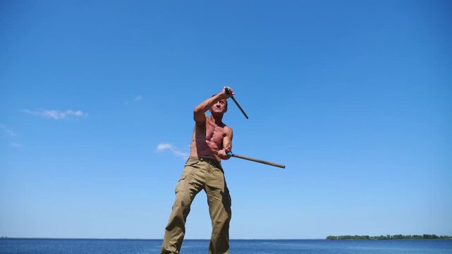 Older sportsman demonstrating wing chun kung fu against blue sea at background. Adult male athlete showing fighting techniques with wooden sticks at sunny summer day. Old man training martial arts