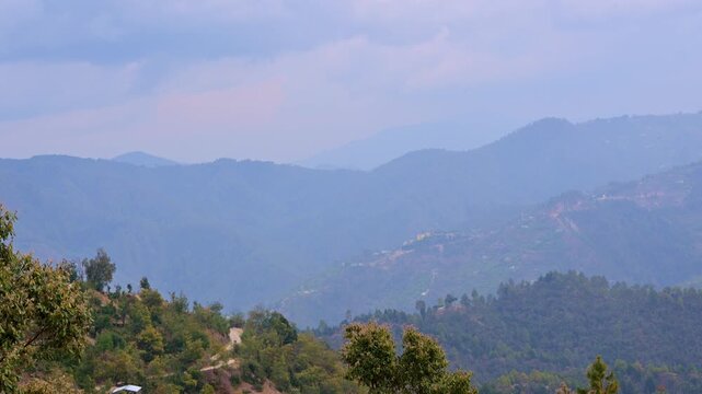 Time-lapse view of Mukteshwar, Uttarakhand, India, capturing layered mountain ranges fading into bluish mist with soft sun rays piercing through clouds, illuminating scattered hillside homes.