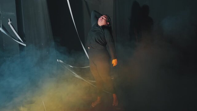 A female contemporary dancer performs dynamic, emotional movements in a dark, fog-filled studio. Heavy smoke and dramatic lighting highlight her expressive body language and artistic passion.