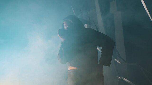 A woman bends forward, catching her breath in a dark, fog-filled room. The heavy smoke and dramatic lighting emphasize her physical and emotional exhaustion after an intense performance.