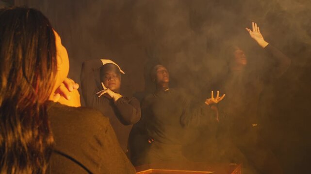 A group of performers in dark clothing execute abstract contemporary dance moves in a room filled with golden smoke. One woman watches from the foreground in this theatrical art piece.