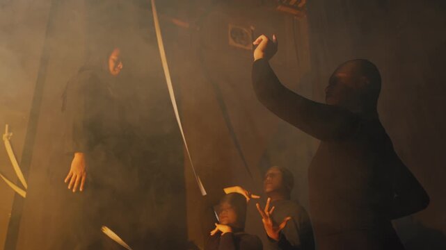 A diverse group of performers in dark attire hold dynamic, dramatic poses on and around a stage prop. Thick yellow smoke and shadows create a compelling artistic visual.