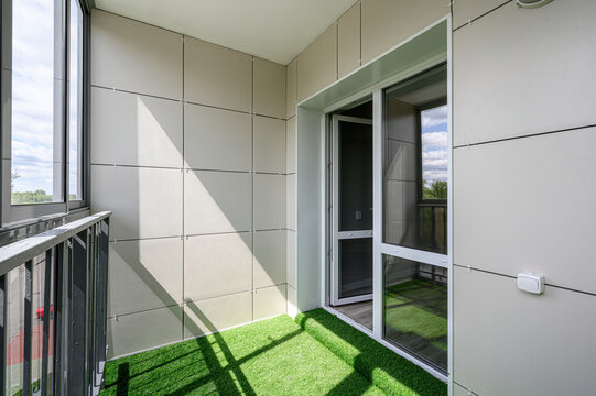 A modern balcony with beige panel walls, artificial green grass flooring, a glass sliding door, metal railing, and large windows offering a view of the sky and trees.