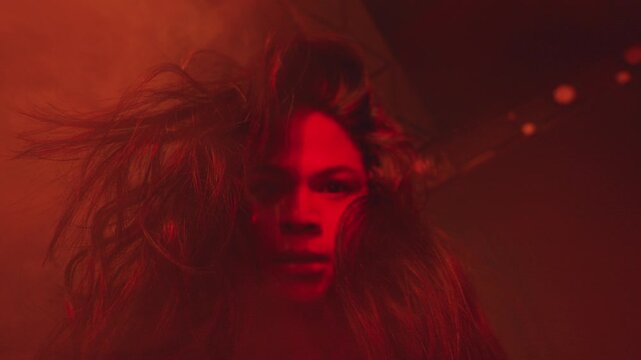 A striking low-angle portrait of a woman with wild hair gazing fiercely into the lens. Engulfed in intense crimson light and thick fog, the shot radiates power, anger, and dominance.