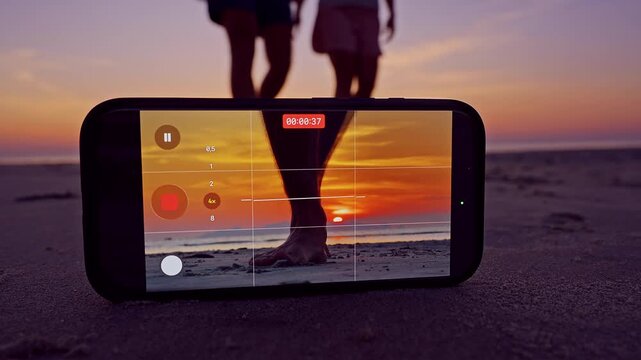 Smartphone on Sand Records Couple Walking Toward Camera at Sunset Beach Screen Shows Grid and Timer While Silhouettes Approach Creating Depth Travel Lifestyle Filming Scene