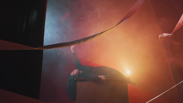 Cinematic wide shot of a female performer lying on a stage prop, bathed in thick fog and warm lights. She slowly reaches her hand upward in a gesture of longing, despair, or awakening.