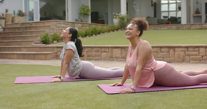 Diverse female friends following cue, holding matching backbends on purple mats on lawn, smiling
