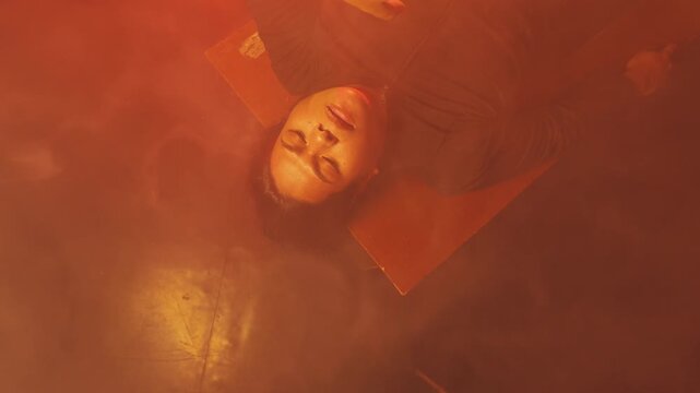 A top-down perspective of a young female lying completely still, enveloped by shifting clouds of thick orange and red haze. Represents a state of limbo, deep sleep, or losing consciousness.