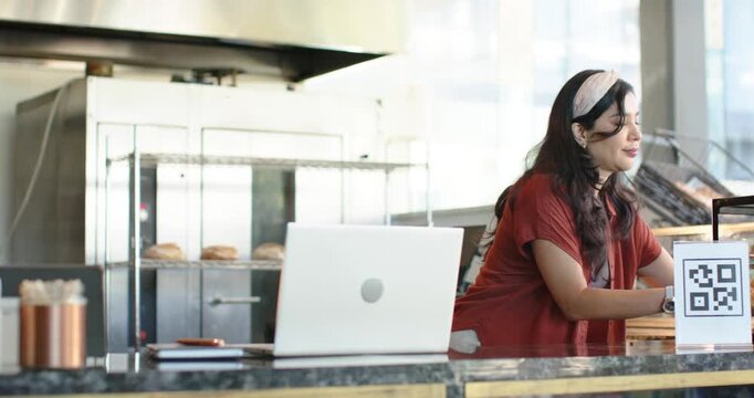 Bakery woman holding pastry tray placing QR sign typing on white laptop handling orders behind till