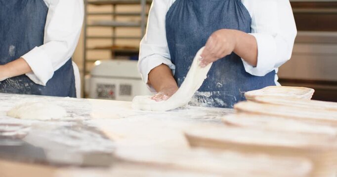 Female bakers, right baker stretching dough on floured bench readying boule for proofing baskets