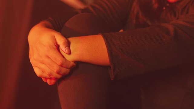 Intimate framing of a hand gripping an arm/leg tightly. The deep red lighting and tense body language highlight a moment of intense anxiety, self-preservation, or emotional pain.