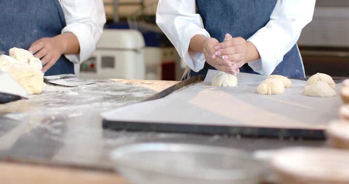 Diverse female bakers left portioning dough with scraper, right rolling rounds for baking at bench
