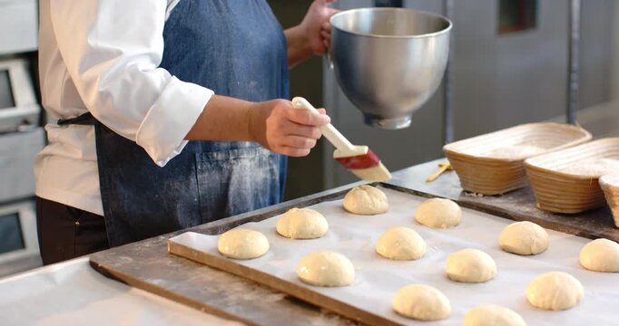 African American woman baker glazing rolls with red-brush at bench dipping into bowl to refill