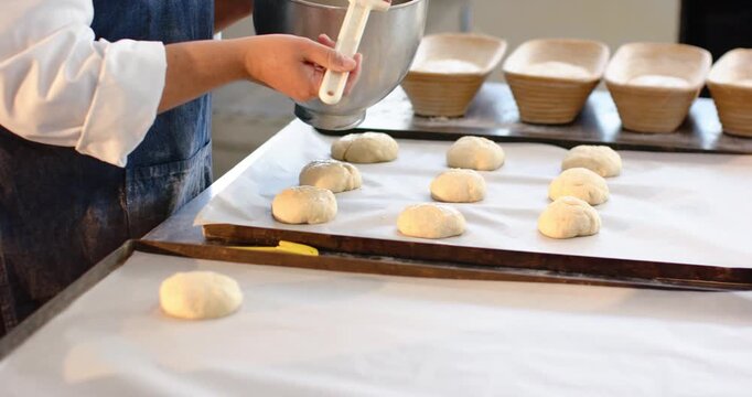 African-American woman in apron tilting bowl and brushing buns for baking at bakery, copy space