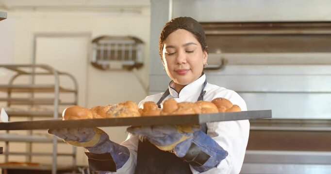 African American woman baker in uniform holding tray, smelling aroma of rolls in commercial bakery