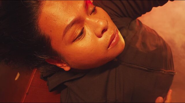 A macro view of a peaceful yet melancholic woman lying down with her eyes closed. Her hand rests gently over her heart in a soothing gesture, bathed in warm, crimson lighting.