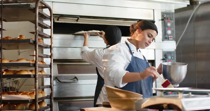 Front baker dipping brush and glazing rolls while rear baker opening oven inserting tray for baking