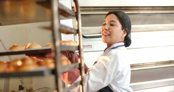 Female baker wearing coat gloves pulling buns from rack at bakery, cooling and high-fiving coworker