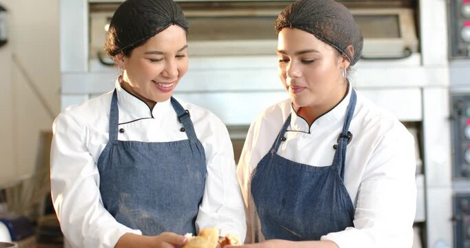 Diverse female bakers in chef jackets in bakery, left presenting roll, right tasting for quality