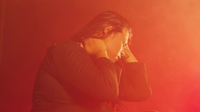 A stressed woman looks up in despair while holding her head, bathed in intense red light and fog. Represents anxiety, feeling trapped, or experiencing an overwhelming sensory overload.