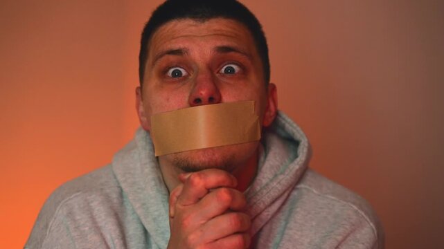 A terrified kidnapped man with his mouth covered with paper tape begs for mercy, the light from a police car illuminates the room and the man,