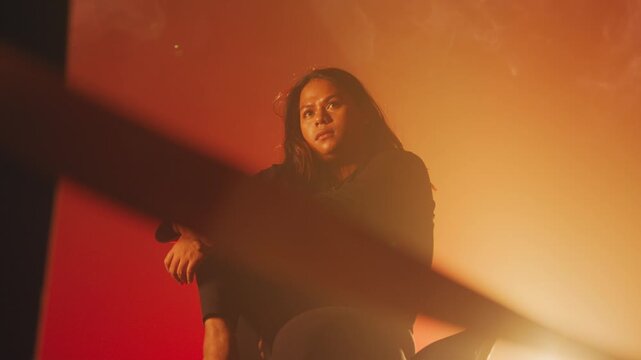 A serious, mysterious person sits in a moody orange and red lit space, looking through foreground framing. Evokes feelings of imprisonment, urban isolation, or deep contemplation.
