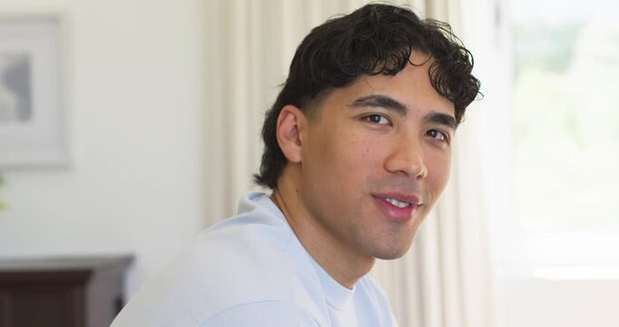 Asian man 20s turning head toward camera shifting expression near bright window wearing blue shirt