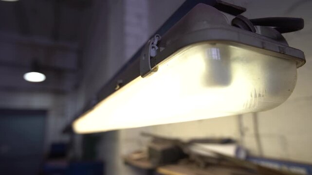 A fluorescent light fixture hangs from the ceiling in a workshop. The light is on and brightens the space. Tools and equipment are visible on a nearby workbench.