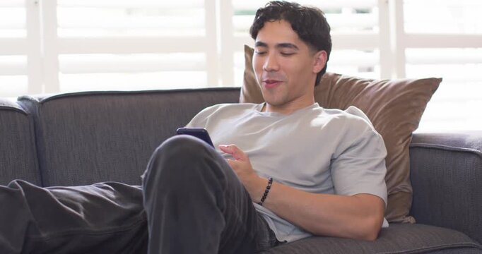 Asian man reclining on dark gray couch, holding smartphone, laughing and wearing beaded bracelet