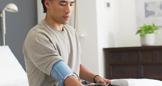 Adult Asian man on bed looking up adjusting blood pressure cuff and placing monitor for reading