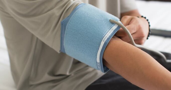Man on bed inserting left arm into blue cuff, fastening Velcro, routing tubing for health check